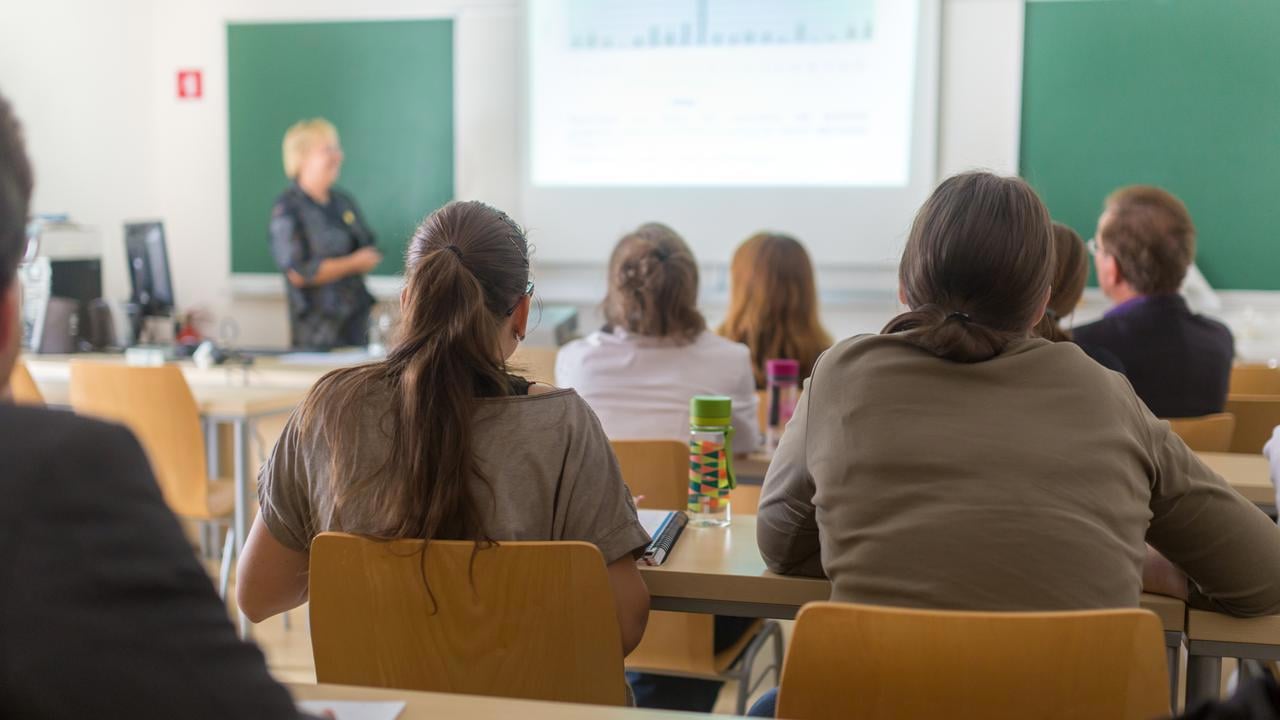 F&ouml;rel&auml;sningssal p&aring; universitet med studenter i f&ouml;rgrunden och en l&auml;rare vid tavlan.