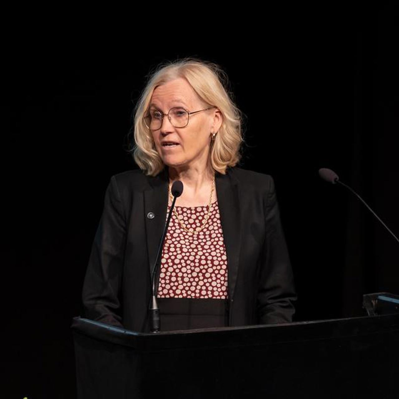 En kvinna st&aring;r p&aring; ett podium framf&ouml;r en mikrofon.