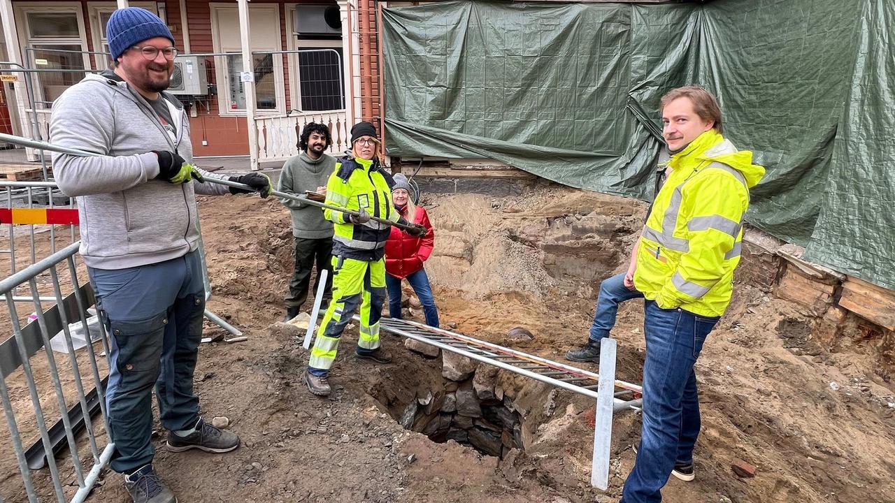 Foto av fem personer i varselkl&auml;der st&aring;r runt en stenlagd gammal brunn p&aring; en byggarbetsplats.