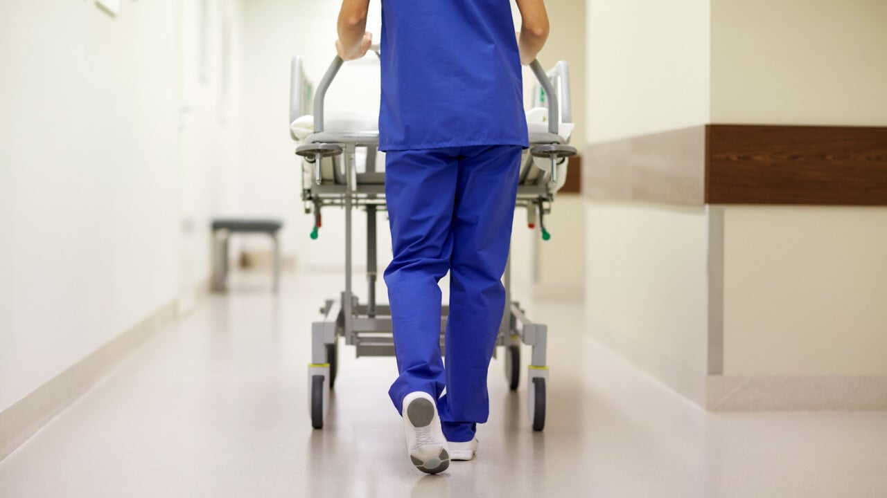 V&aring;rdpersonal skjuter en s&auml;ng p&aring; hjul med patient i en sjukhuskorridor.