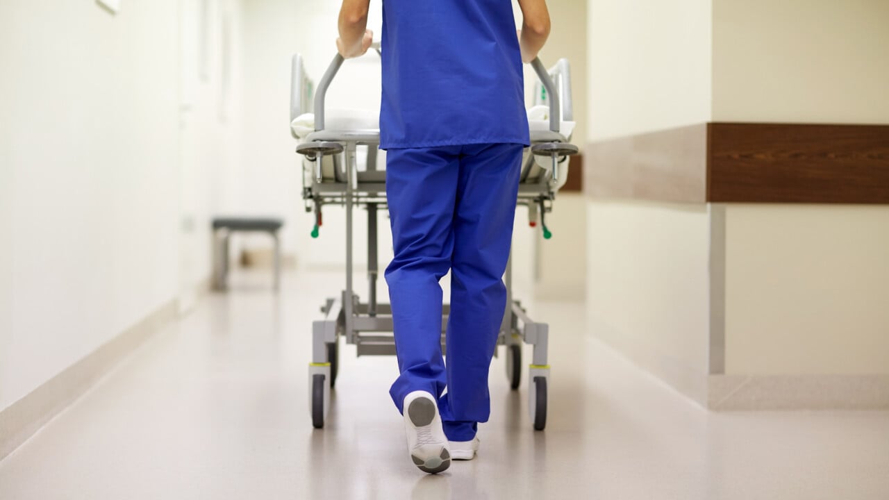 V&aring;rdpersonal skjuter en s&auml;ng p&aring; hjul med patient i en sjukhuskorridor.