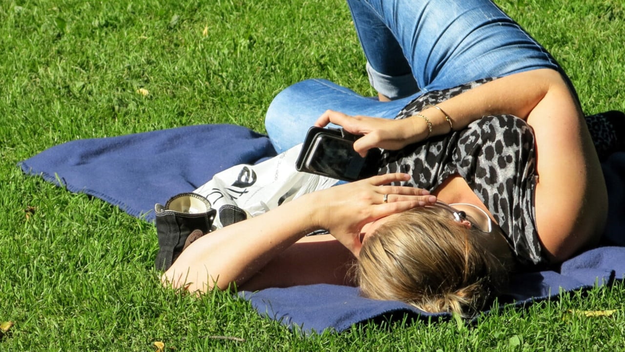 Kvinna med mobiltelefon p&aring; ligger p&aring; filt p&aring; gr&auml;smatta och surfar.