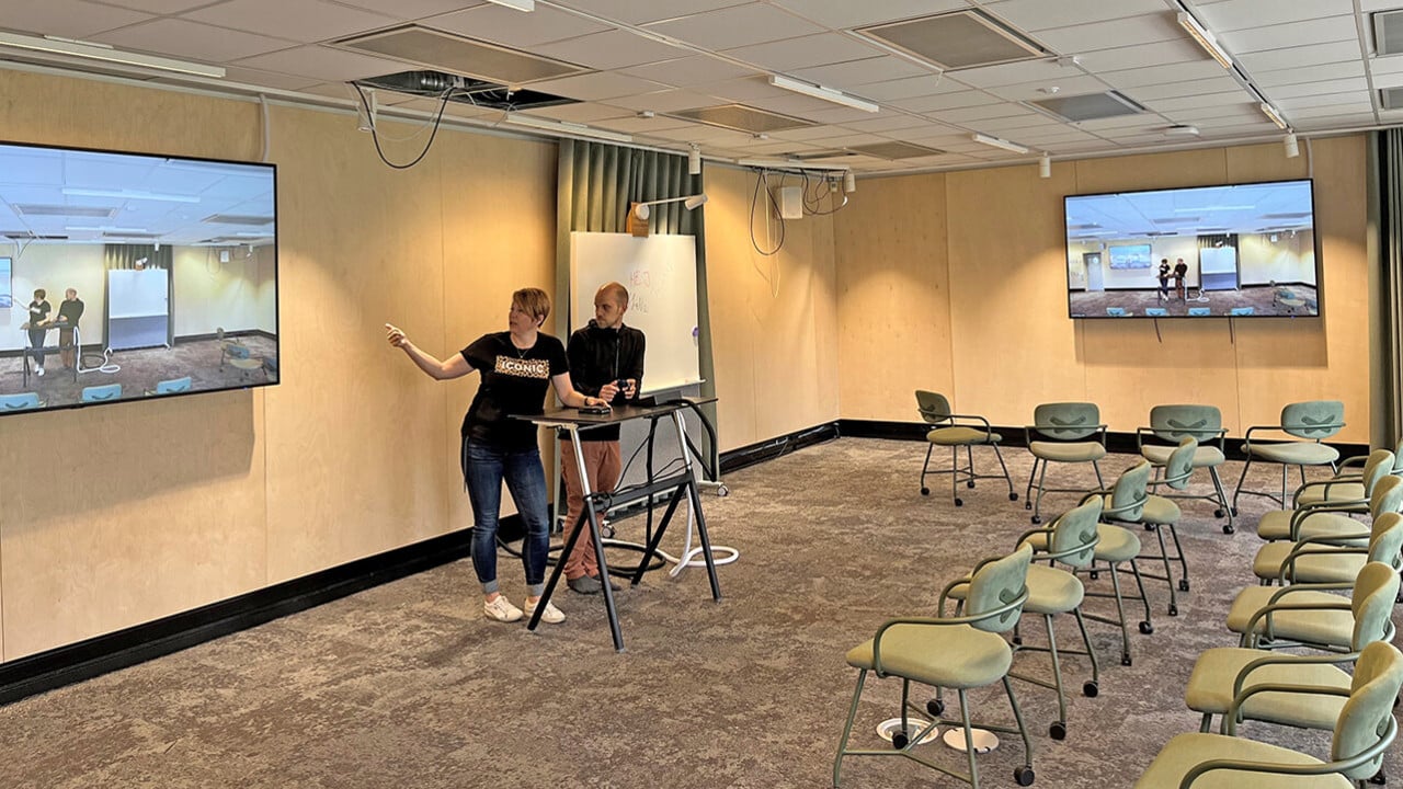 &Aring;se Tieva och Jonas Lindholm, UPL, stand at a podium and look at themselves on a large screen to their left. On their right is a whiteboard and another large screen showing the same image.