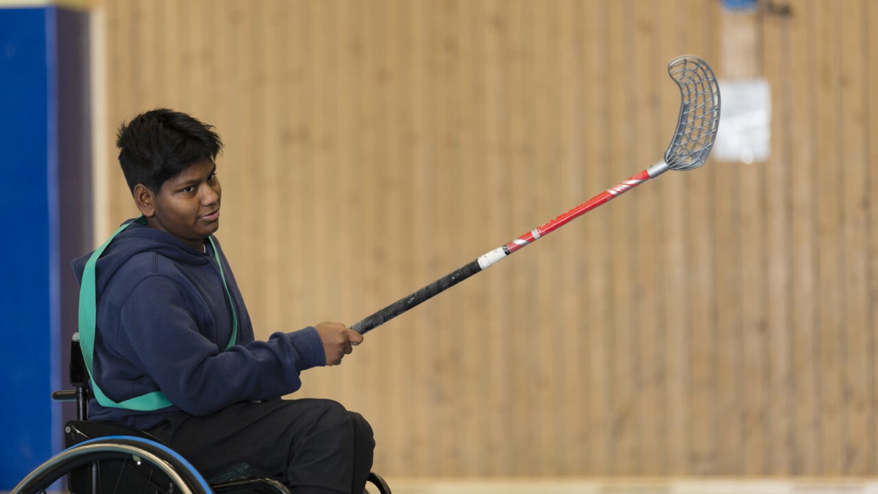 Bild p&aring; kille i rullstol som med innebandyklubba i handen skjuter iv&auml;g en boll.