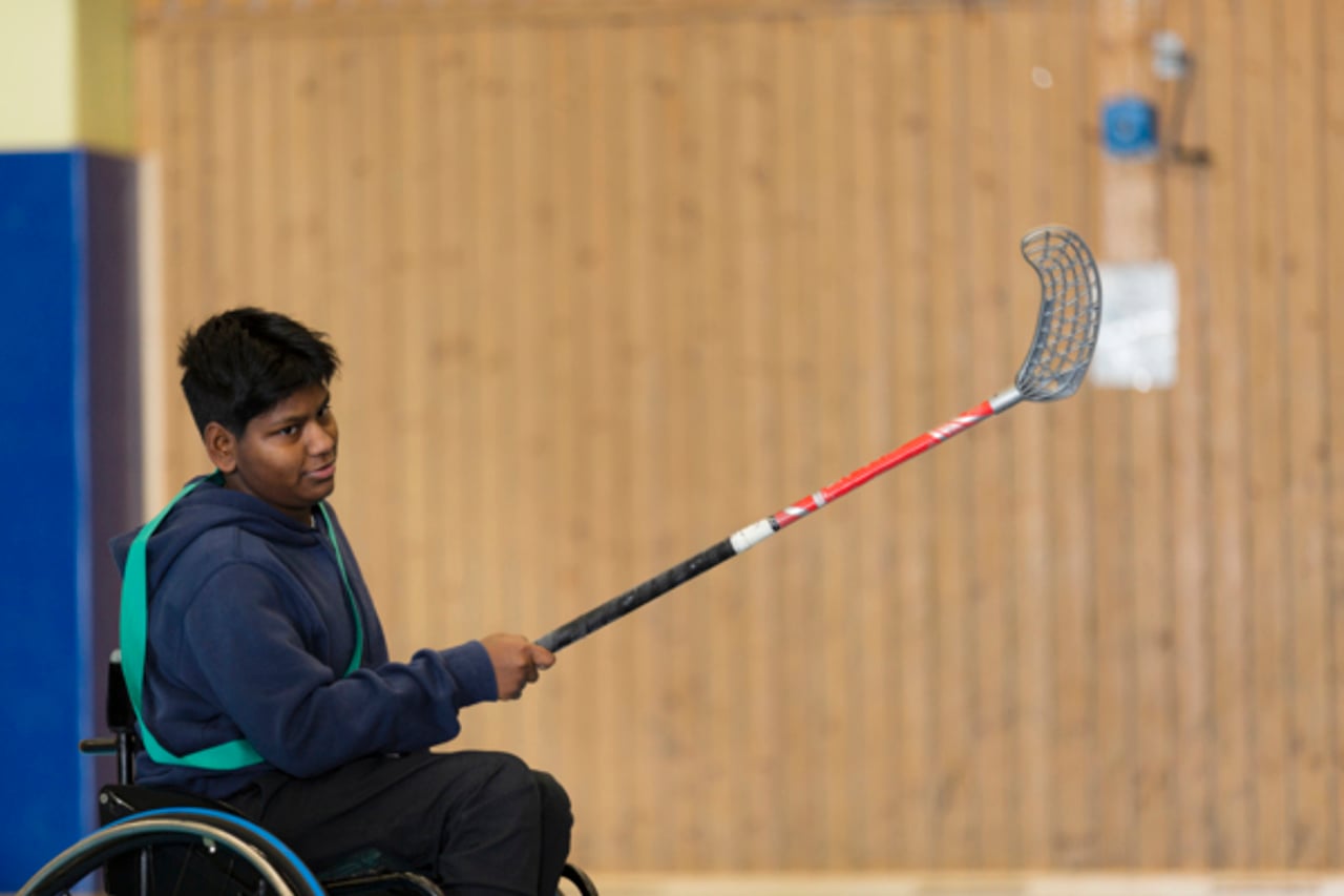 Kille i rullstol med innebandyklubba i handen skjuter iv&auml;g en boll.
