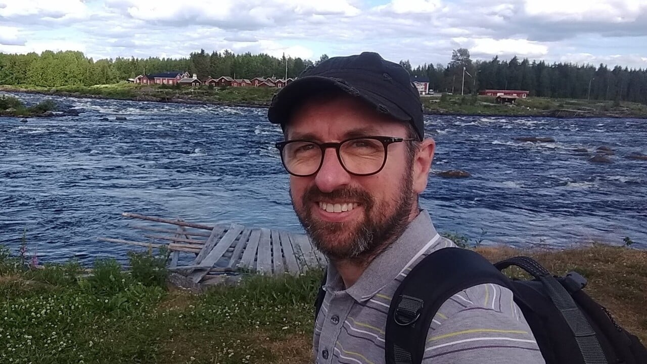 Stuart Wales st&aring;r framf&ouml;r en &auml;lv. Stugor i bakgrunden.