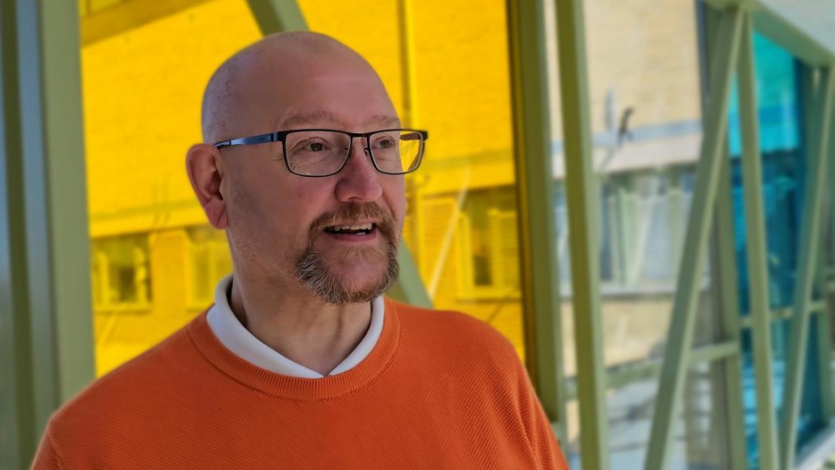 Man utan hår, med svarta glasögon, en orangetröja med vid skjorta under, som står framför ett fönster. 