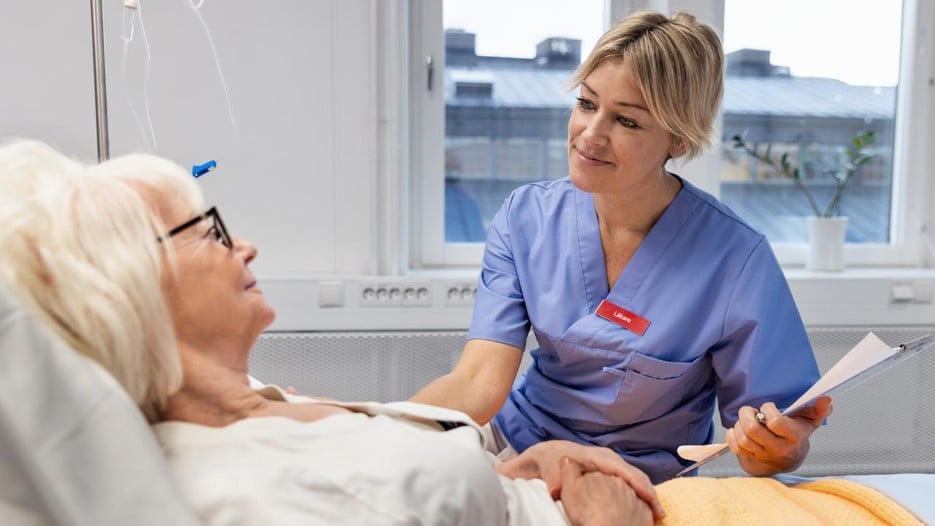 A nurse is sitting next to a bed where an elderly woman is lying