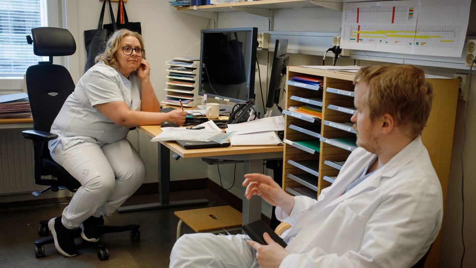 Forskningssjuksköterskan och studiekoordinatorn Nina Sundqvist sitter vid sin dator och tar anteckningar. Hon tittar mot läkaren och doktoranden Ivar Winroth som sitter i en besöksstol i förgrunden och pratar med henne.