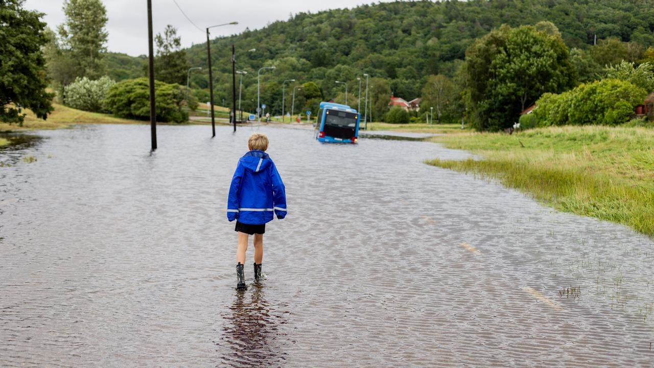 Ett barn står vänd mot en översvämmad bilväg. Längre fram syns en buss.