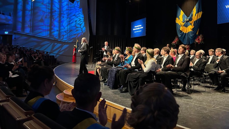 People sit on chairs on a stage, and one man stands in the front and recieve a diploma.