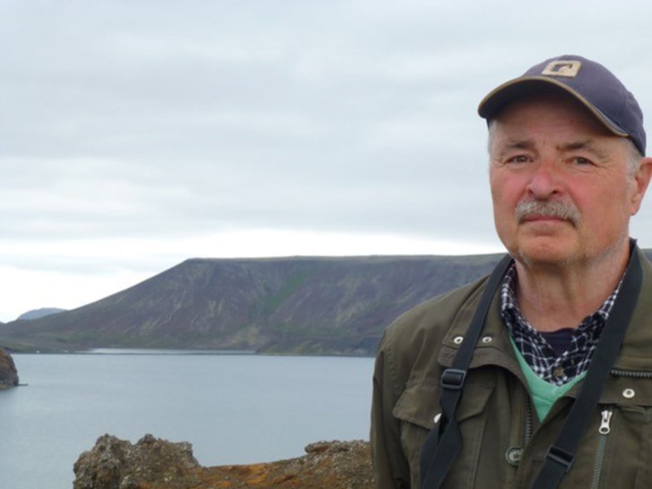 Bild på Lennart Bondesson som utomhus med en sjö och berg i bakgrunden. Han bär en keps och en grön jacka och ser direkt in i kameran.