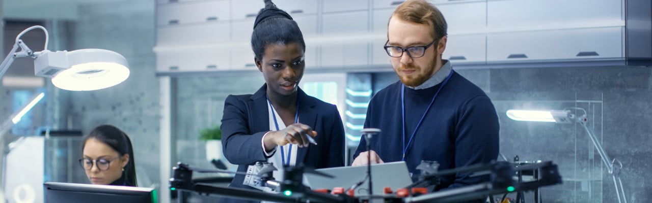 Caucasian Male and Black Female Engineers Working on a Drone Project with Help of Laptop and Taking Notes. He Works in a Bright Modern High-Tech Laboratory.