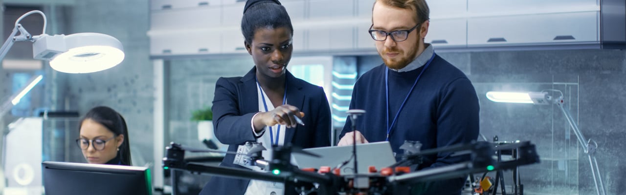 Caucasian Male and Black Female Engineers Working on a Drone Project with Help of Laptop and Taking Notes. He Works in a Bright Modern High-Tech Laboratory.