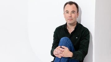Marcus Gustafsson sitter med knät uppdraget och armarna runt benet, han tittar rakt in i kameran. Han har jeans och en svart skjorta på sig. 