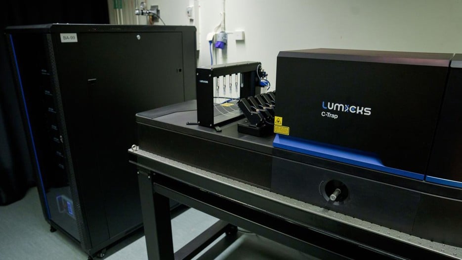 A black high box is standing in the back and a black table with a stand with capillaries and another black box in front on which "Lumicks C-Trap" is written on.