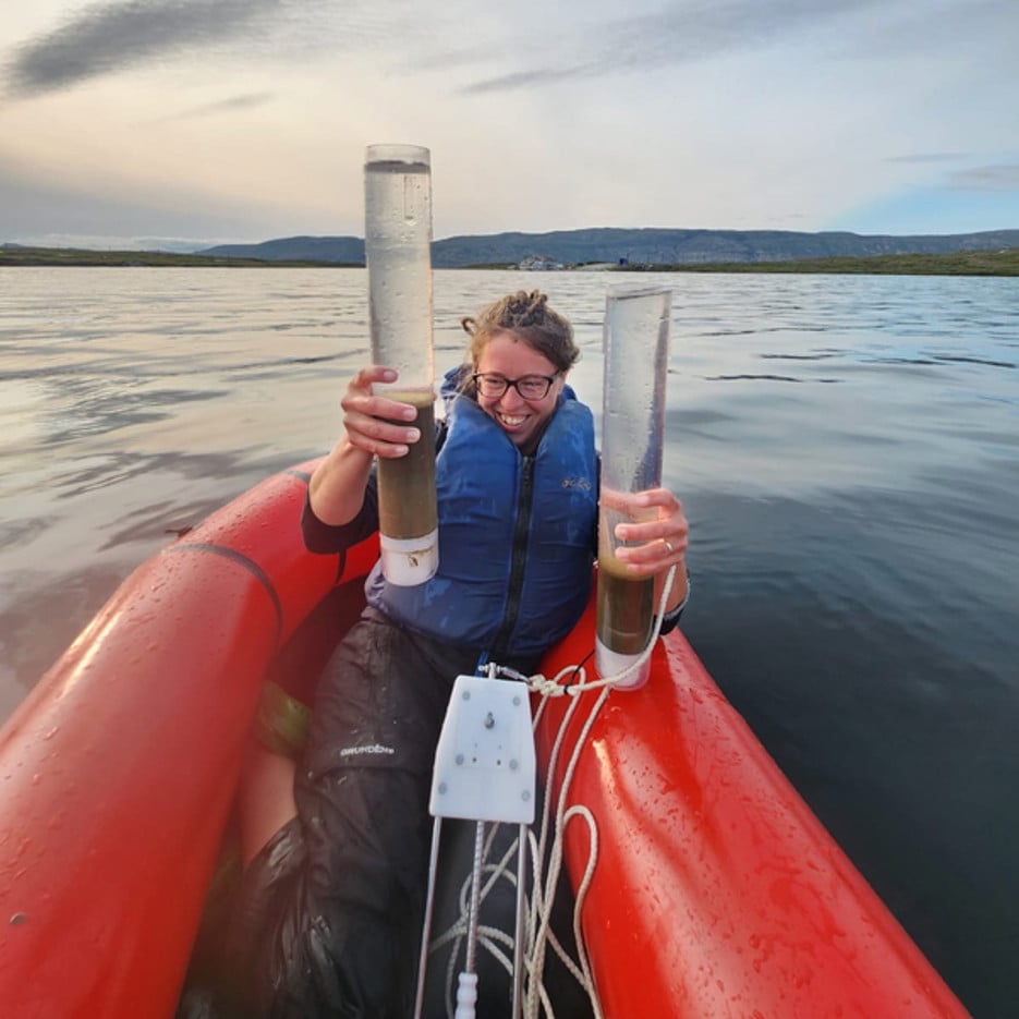 Václava Hazuková samlar in sedimentprovet från en arktisk sjö. Hon sitter i en liten röd gummibåt och håller upp två stora cylindriska behållare med vatten och sediment. 