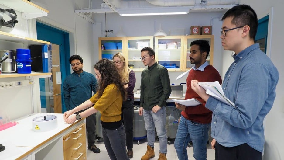 Students standing around the teacher in a lab, the teacher shows different tools to be used. Some students take notes. 