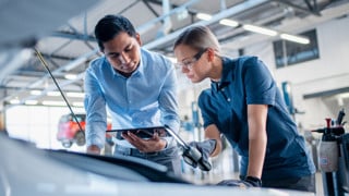 Instructor with a Tablet Computer is Giving a Task for a Future Mechanic. Female Student Inspects the Car Engine. Assistant is Checking the Cause of a Breakdown in the Vehicle in a Car Service.