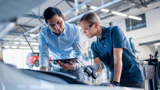 Instructor with a Tablet Computer is Giving a Task for a Future Mechanic. Female Student Inspects the Car Engine. Assistant is Checking the Cause of a Breakdown in the Vehicle in a Car Service.