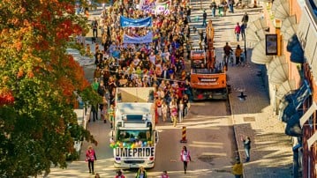Drönarbild av en parad av människor som går i Pride-tåget i Umeå 2024, med regnbågsflaggor och banderoller. 