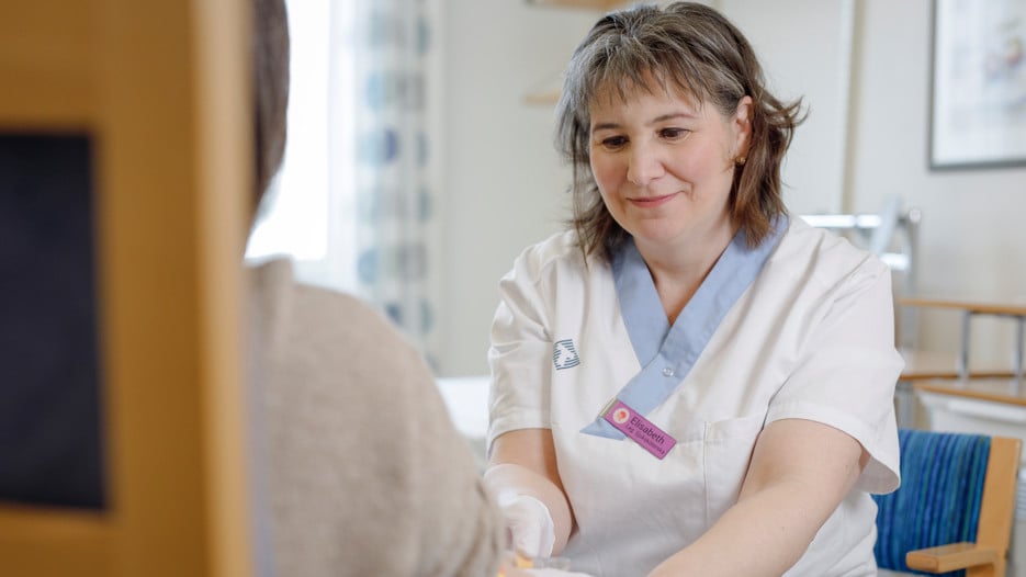 A person i seen sitting with their back against the camera, holding their arm out. Sitting across from them is Elisabeth Müller Granberg, about to take a blood sample. 