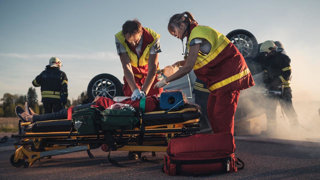 On the Car Crash Traffic Accident Scene: Paramedics Saving Life of a Female Victim who is Lying on Stretchers. They Apply Oxygen Mask, Do Cardiopulmonary Resuscitation / CPR and Perform First Aid