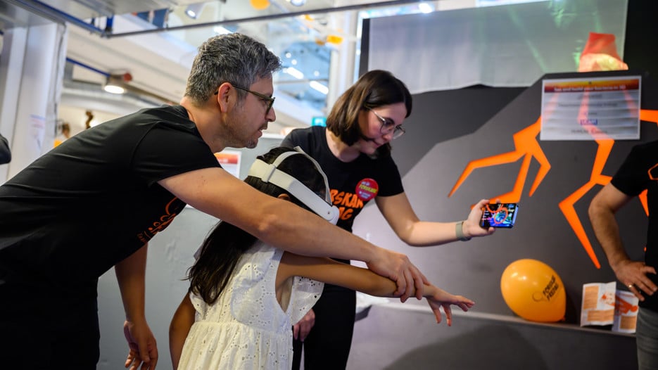 A man and a woman guide a child in a white dress with a white VR headset on