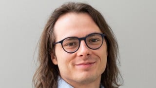 A man with long hair wearing glasses and a blue shirt. 