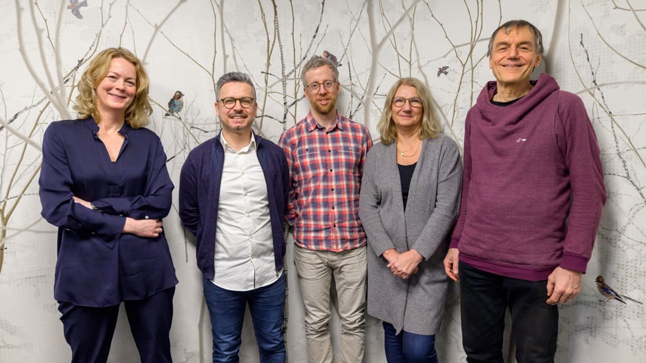 Five people standing in front of a wallpaper of a forest with birds