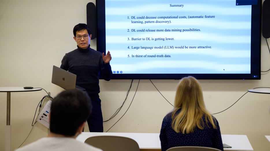Jian-Feng Mao stands in front and to the side of a screen showcasing a summary slide. He is holding his laptop in one hand and gestures with the other.