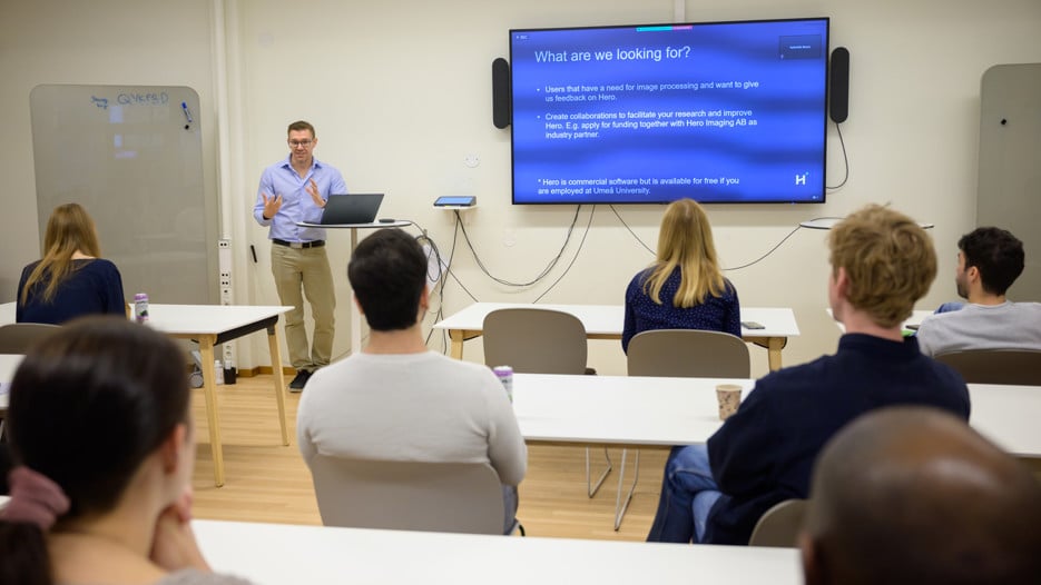 A group of researchers are seated at white tables with their backs to the camera. They face Anders Garpebring who stands by a round table with his laptop. The screen next to him shows a slide that says "What am I looking for?" followed by more text.