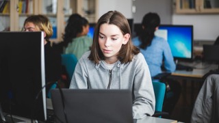Confident female student using laptop at desk against teacher and friends sitting in computer lab