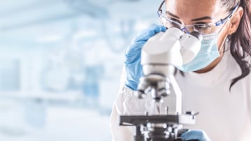 Female lab technician in protective glasses, gloves and face mask sits next to a microscope in laboratory.