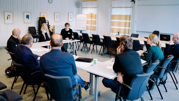 Åtta personer sitter vid bord formade som en hästsko i ett konferensrum.