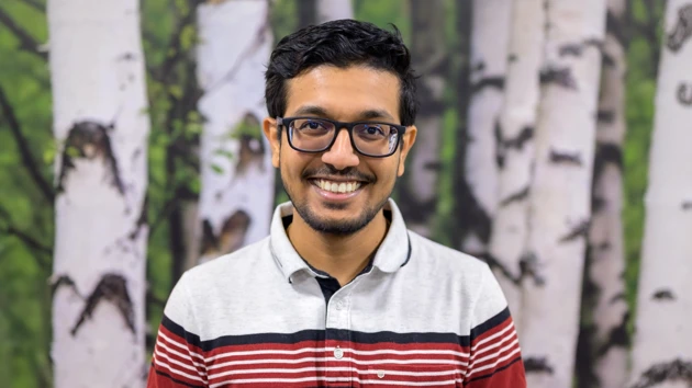 Man with glasses and a red and white shirt sits in front of a birch tree background and smiles. 