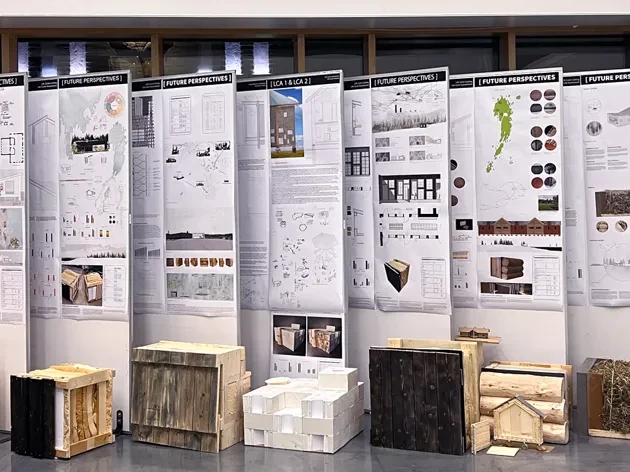 Exhibition in Umeå School of Architecture's entrance with built objects and posters, dark outside