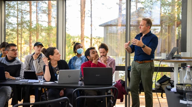 En grupp studenter sitter vid bord med laptops uppslagna och lyssnar på en man som står och talar samtidigt som han gestikulerar med armarna. De befinner sig i ett glasat rum, utanför syns träd och ett annat trähus. 