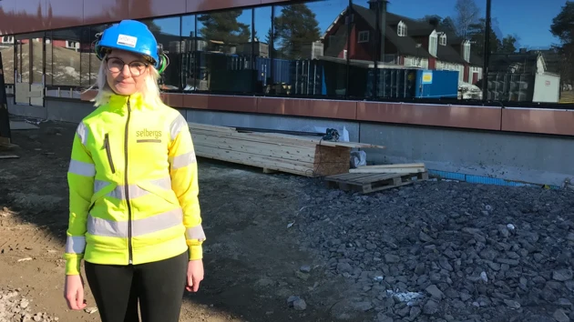 En kvinna i varselkläder och blå bygghjälm tittar mot kameran på en byggarbetsplats.