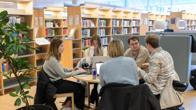 Three people sitting at a table in the library studying. Around the table are several bookshelves with books.