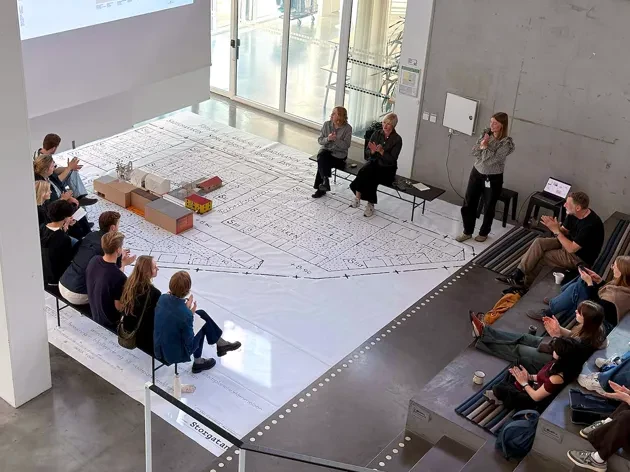 Lecture in big open space with lecturer's, people watching and a big urban plan on the floor