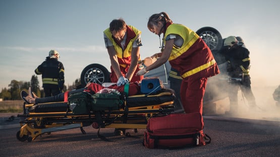 On the Car Crash Traffic Accident Scene: Paramedics Saving Life of a Female Victim who is Lying on Stretchers. They Apply Oxygen Mask, Do Cardiopulmonary Resuscitation / CPR and Perform First Aid