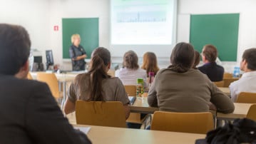Föreläsningssal på universitet med studenter i förgrunden och en lärare vid tavlan.