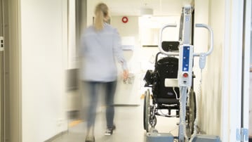 Korridor i vårdhem med rullstol och andra hjälpmedel samt personal som skyndar fram.