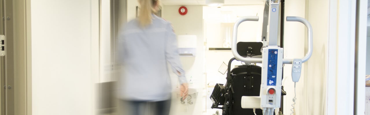 Korridor i vårdhem med rullstol och andra hjälpmedel samt personal som skyndar fram.