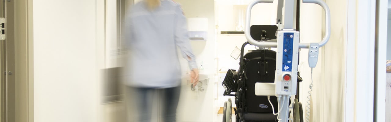 Korridor i vårdhem med rullstol och andra hjälpmedel samt personal som skyndar fram.