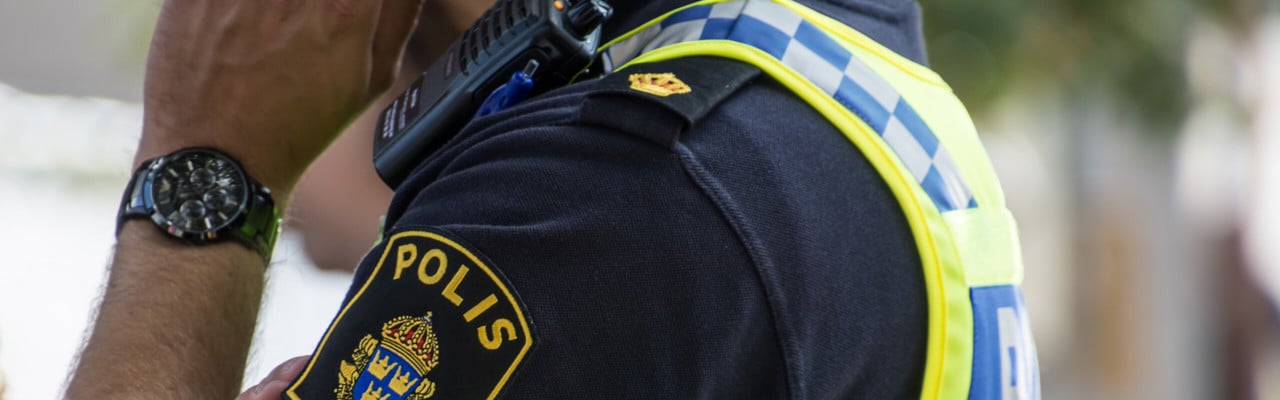 Detalj polis med uniform som pratar i mobil. Man ser arm och tröja med polisens emblem.
