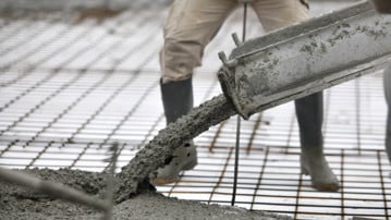 Detalj från ett bygge där betonggjutning pågår. Cement rinner ner på armeringsjärn och arbetare skymtar i bakgrunden.