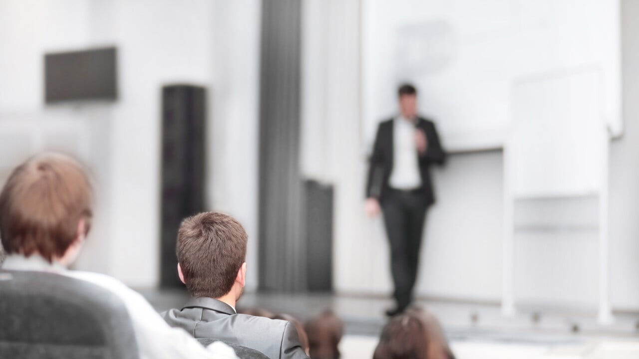 Genrebild på en föreläsare vid tavlan med publik i förgrunden. För att illustrera föreläsning eller seminarium.