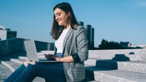 Bilden visar en kvinna som sitter och arbetar med en laptop, utomhus på en trappa i solen.